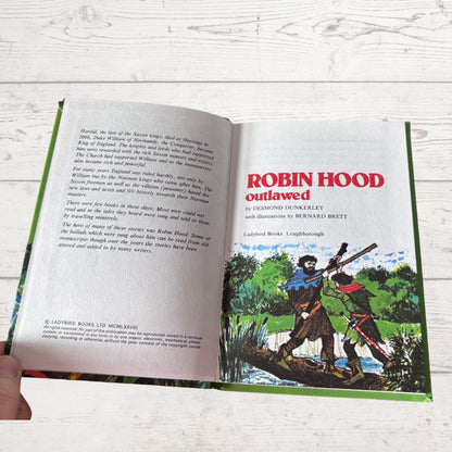An open hardcover book titled 'Robin Hood outlawed' showing printed text on the left page and a colorful illustration on the right page, photographed on a white wood surface with a hand holding the lower left corner.