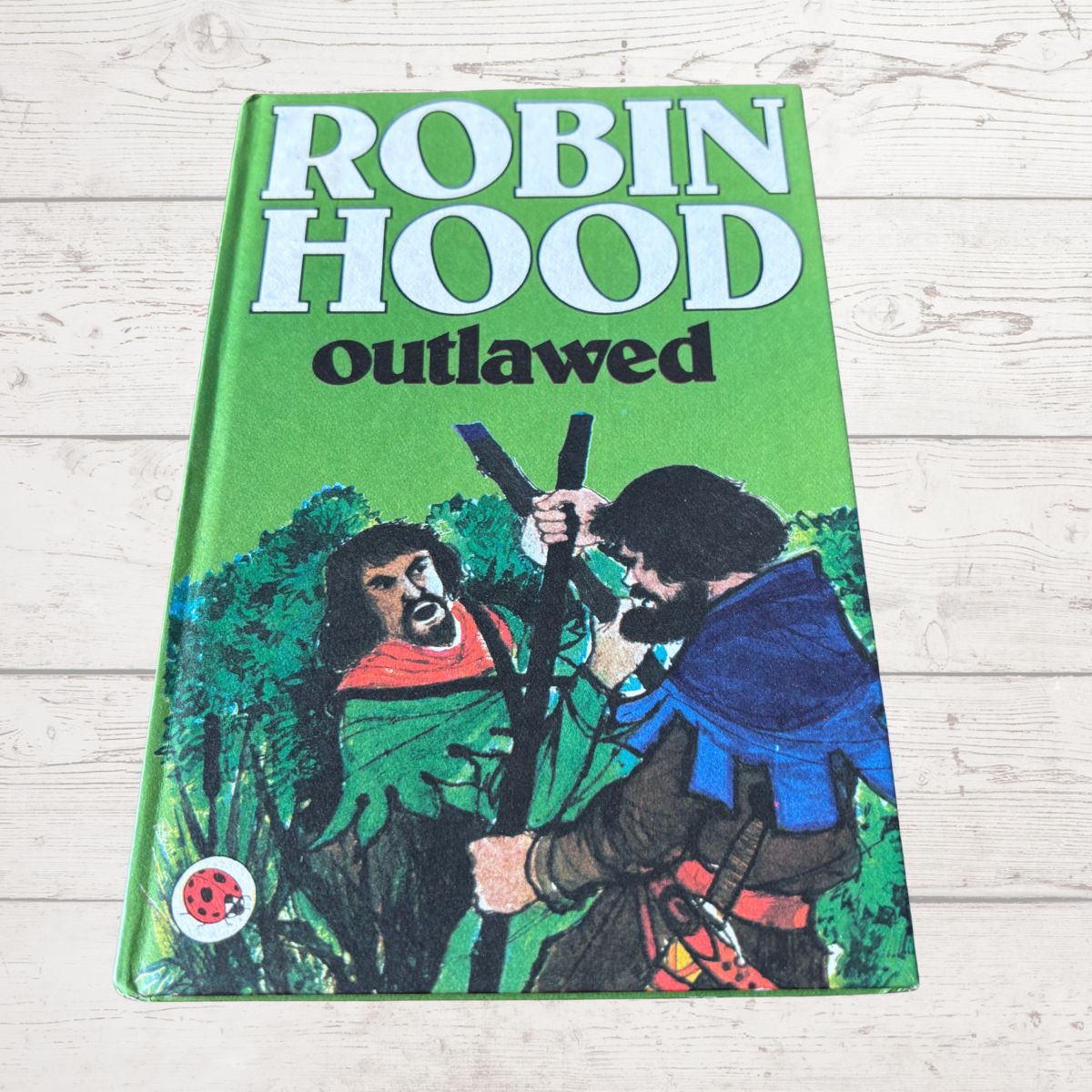 A vintage hardcover book of 'Robin Hood Outlawed' lying flat on a light-colored wooden surface. The book cover features a bright green background with bold white and black title text, and an illustration of two figures in medieval clothing.