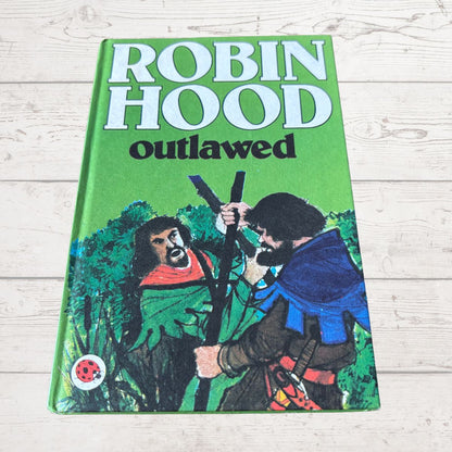 A vintage hardcover book of 'Robin Hood Outlawed' lying flat on a light-colored wooden surface. The book cover features a bright green background with bold white and black title text, and an illustration of two figures in medieval clothing.