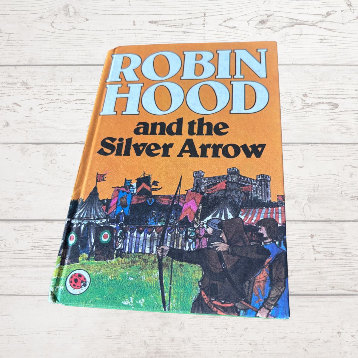A vintage paperback book of Robin Hood and the Silver Arrow lying flat on a light-colored wooden surface. The cover features a prominent orange background with the title in bold white and black lettering, and an illustration of medieval figures, tents, and a castle at the bottom.