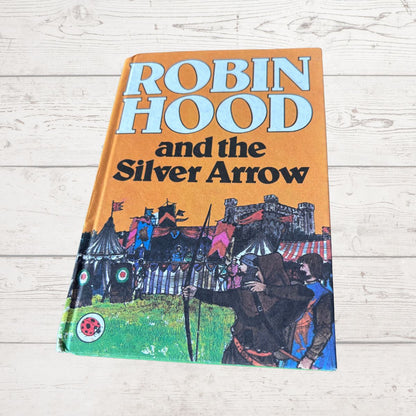 A vintage paperback book of Robin Hood and the Silver Arrow lying flat on a light-colored wooden surface. The cover features a prominent orange background with the title in bold white and black lettering, and an illustration of medieval figures, tents, and a castle at the bottom.