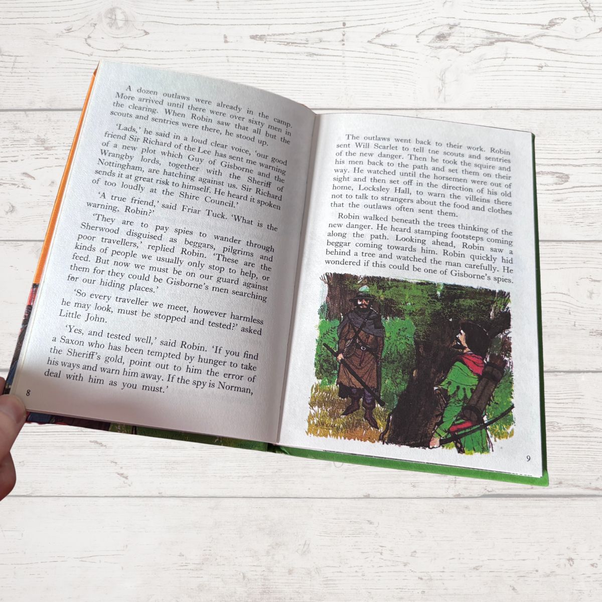 An open children's book lying on a light wood surface, with a hand holding the lower left corner. The book shows printed text pages and a colorful illustration on the right page with a red bird and green foliage.