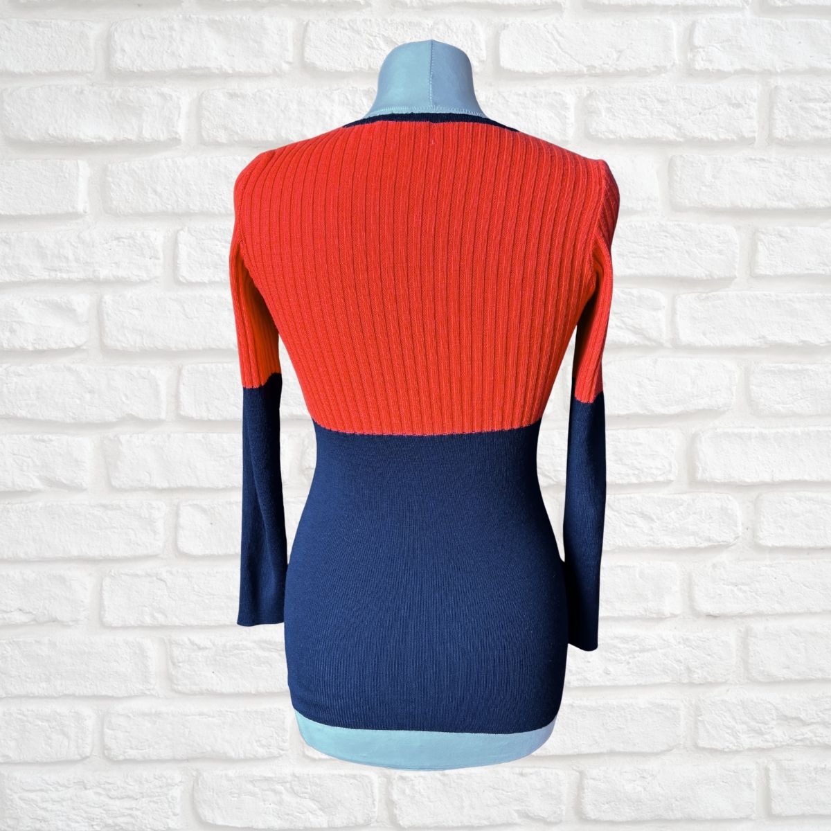 A color-blocked ribbed knit long-sleeve sweater displayed on a mannequin torso against a white brick wall background. The top half is a vibrant red-orange, the bottom half is navy blue, and the sleeves are solid navy blue.