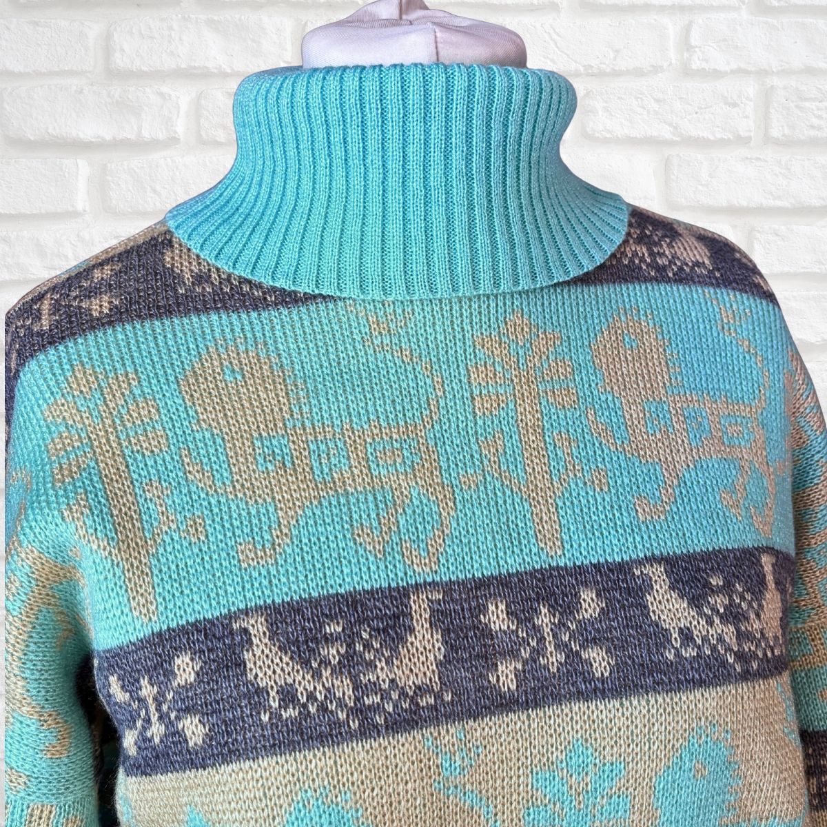 A close-up of a knit turtleneck sweater on a mannequin torso, featuring a turquoise blue base with beige and navy blue Nordic-style patterns including reindeer and snowflakes, set against a white brick wall background.