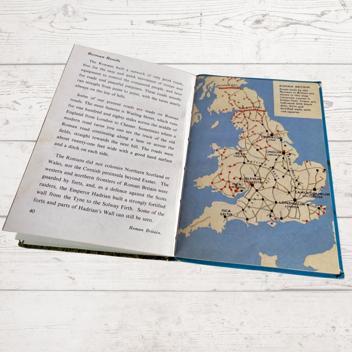 An open vintage book showing a map of Roman Britain and accompanying text on the left page, photographed on a white wood surface.