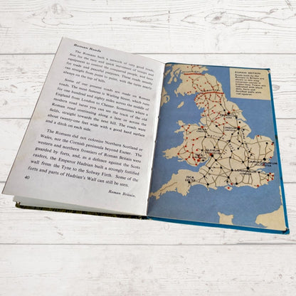 An open vintage book showing a map of Roman Britain and accompanying text on the left page, photographed on a white wood surface.
