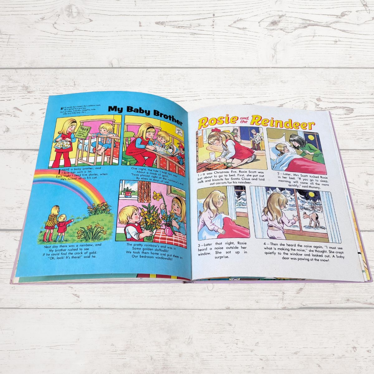 An open children's book spread laid flat on a white wood surface, showing two illustrated pages with colorful comic panels and text. The left page has the title 'My Baby Brother' and the right page has the title 'Rosie the Reindeer'.