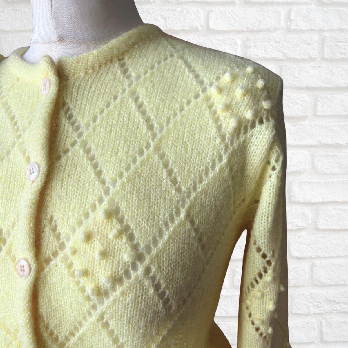Light yellow  knitted cardigan on a mannequin against a white brick wall.
