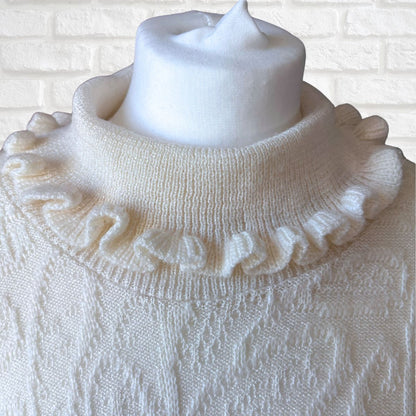 A cream-colored knit sweater with a ruffled mock neck collar, displayed on a white mannequin torso against a white brick wall background.