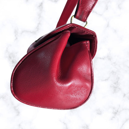 A deep red leather handbag with a curved, teardrop-like silhouette and a matching strap, shown from a top-down angle on a white marble background.