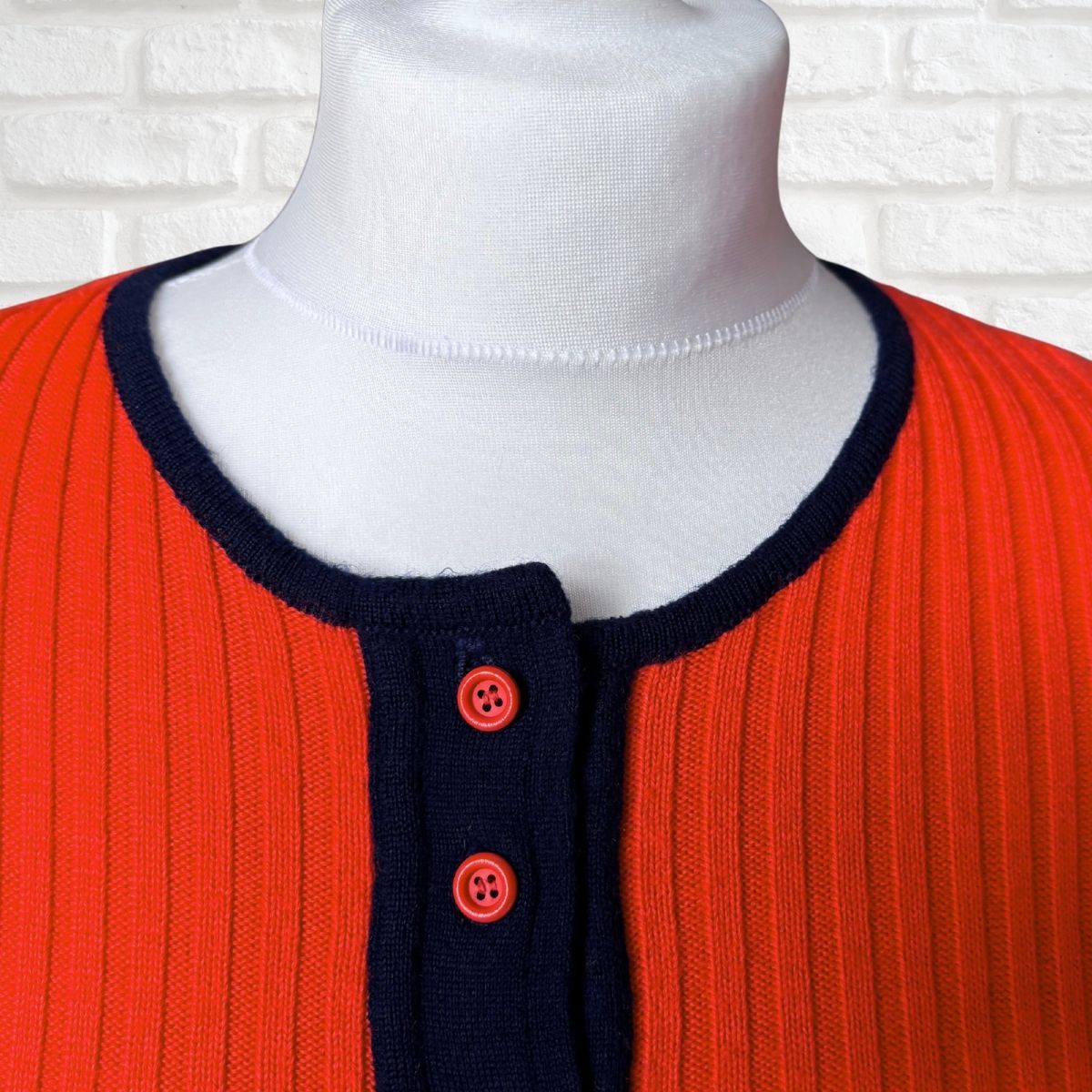 A close-up of a ribbed knit sweater displayed on a white mannequin torso. The cardigan features a vibrant orange-red body with a contrasting dark navy blue ribbed trim around the crew neck and a matching navy button placket with two red buttons.