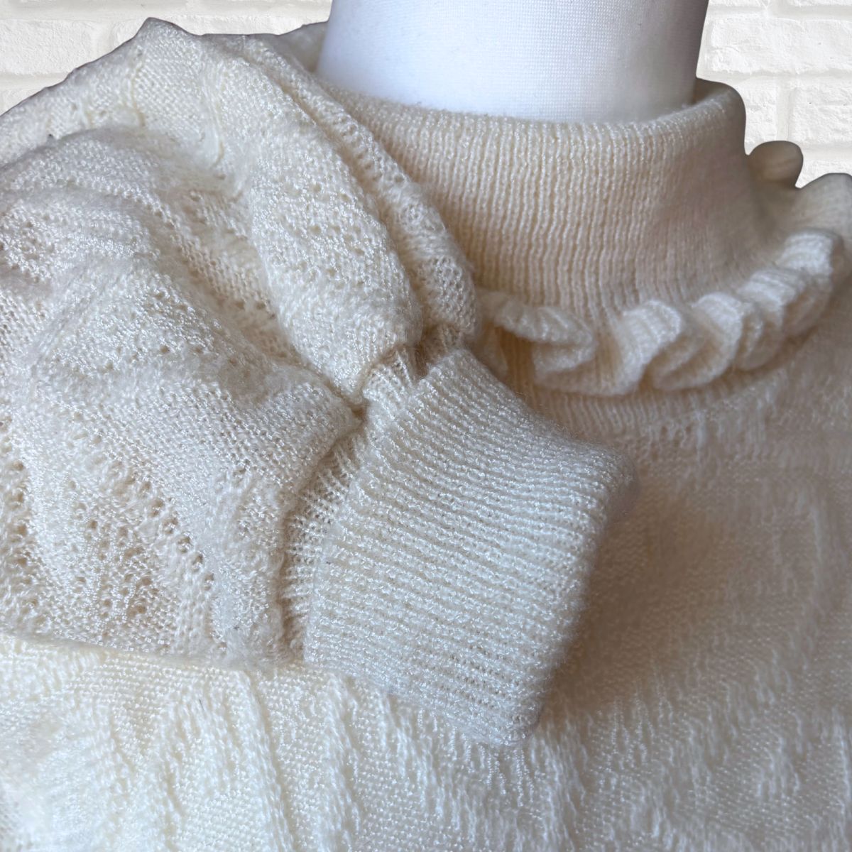 Close-up of a cream-colored knit sweater on a mannequin, showing textured knit patterns, a ruffled mock neck, and a cuffed sleeve.
