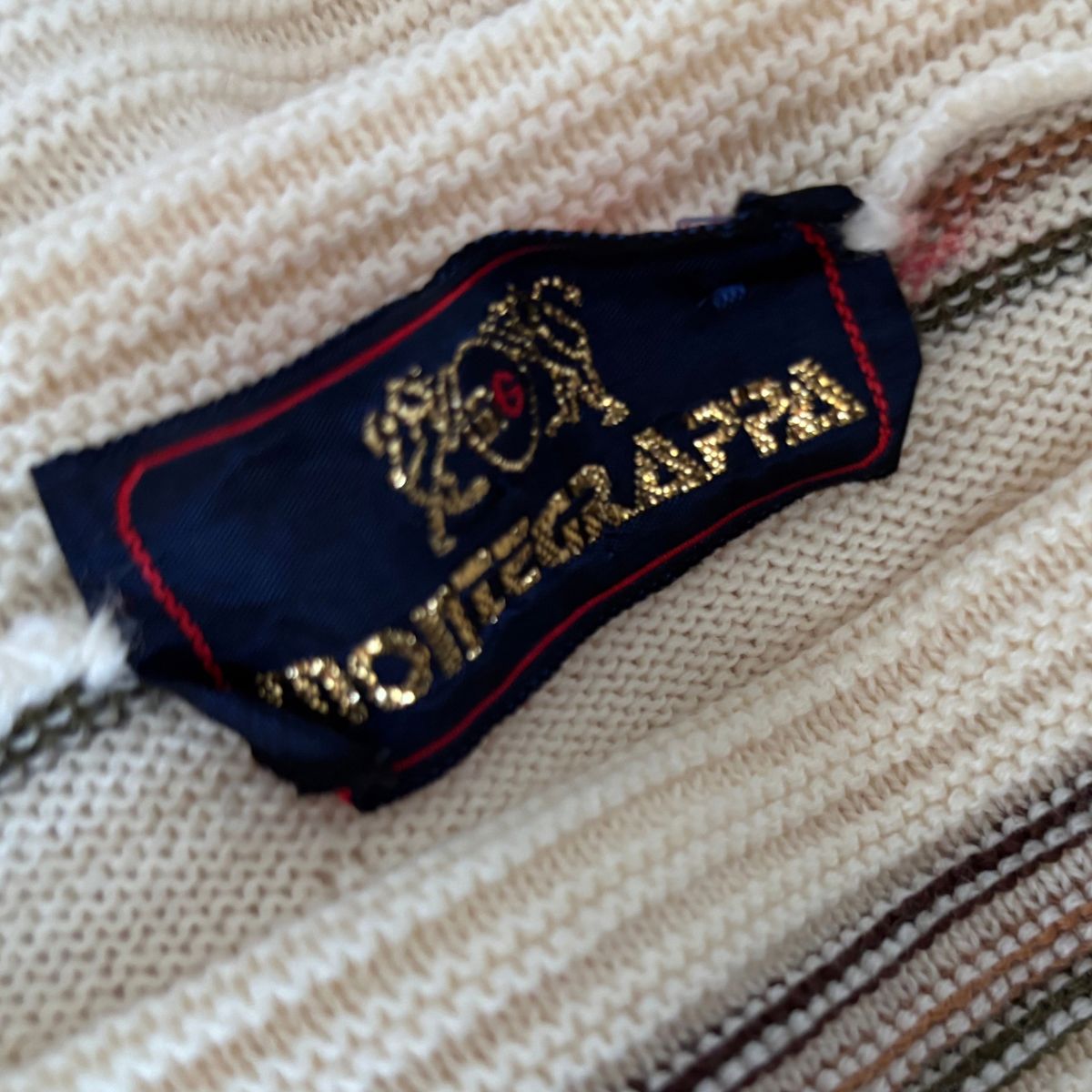 Close-up of a knit sweater tag reading MONTEGRAFA with a gold crest and red trim on a cream-colored ribbed knit fabric.