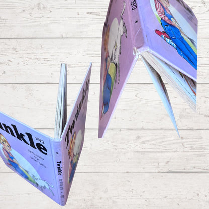 A children's hardcover book titled 'Twinkle' with colorful illustrated pages, shown partially open on a light wood plank background.