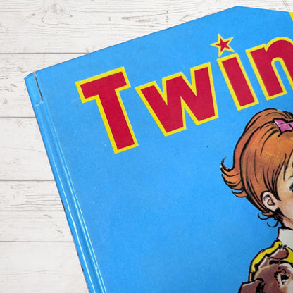 A close-up of a children's book cover with a bright blue background, large red title lettering outlined in yellow, and part of a cartoon girl with brown hair on the right side, placed on a white wood surface.