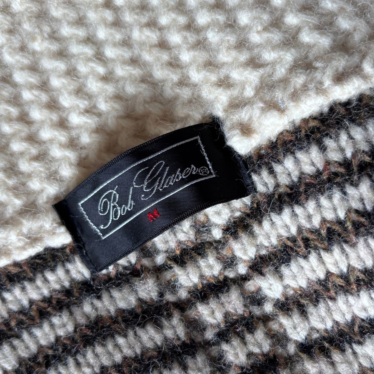 Close-up of a knit textile product showing a black fabric label with white script reading Bob Glaser and a small red 'M' size marking, against cream and black-and-beige striped knit fabric.