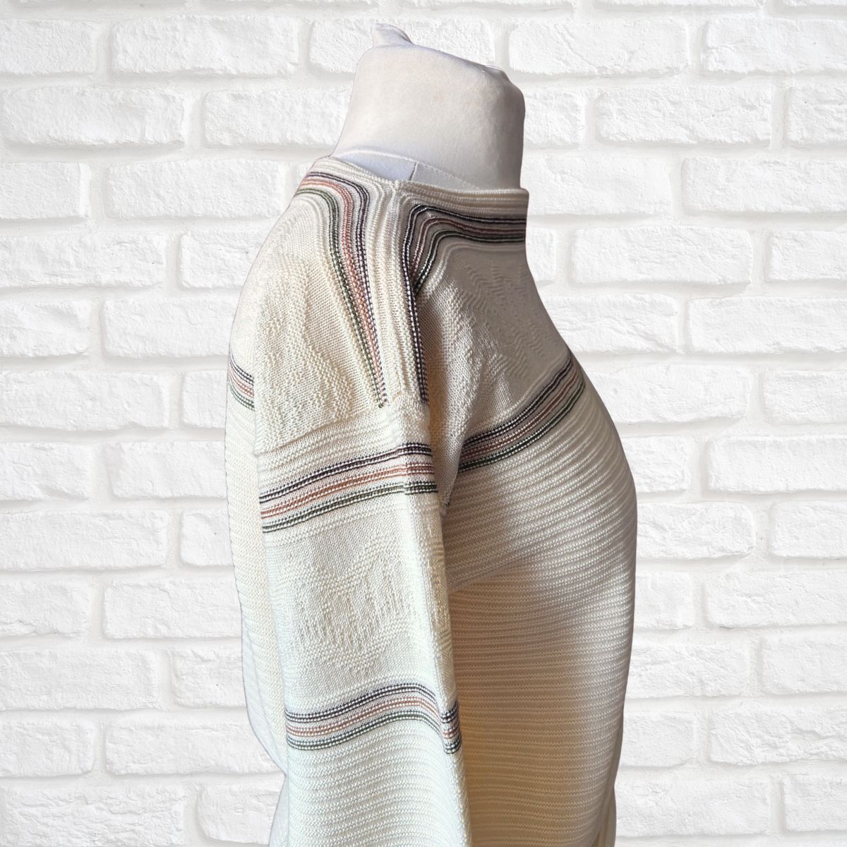 A cream-colored ribbed knit sweater with thin striped accents  on a white mannequin torso against a white brick wall background