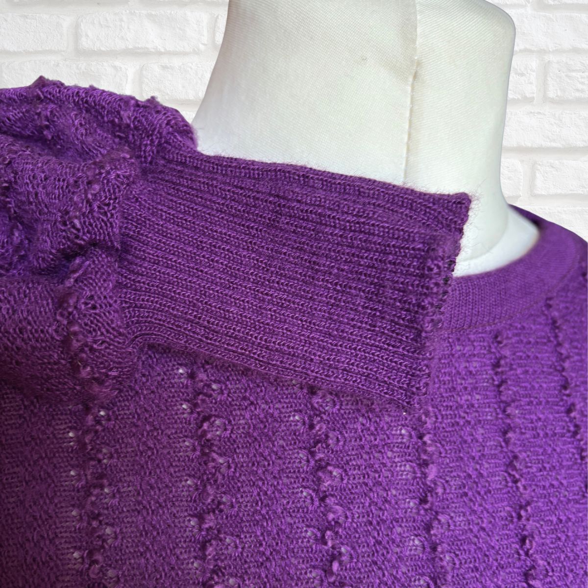 A close-up of a purple knit sweater on a white mannequin torso, showing a textured crew neck and ribbed collar against a white brick wall background.