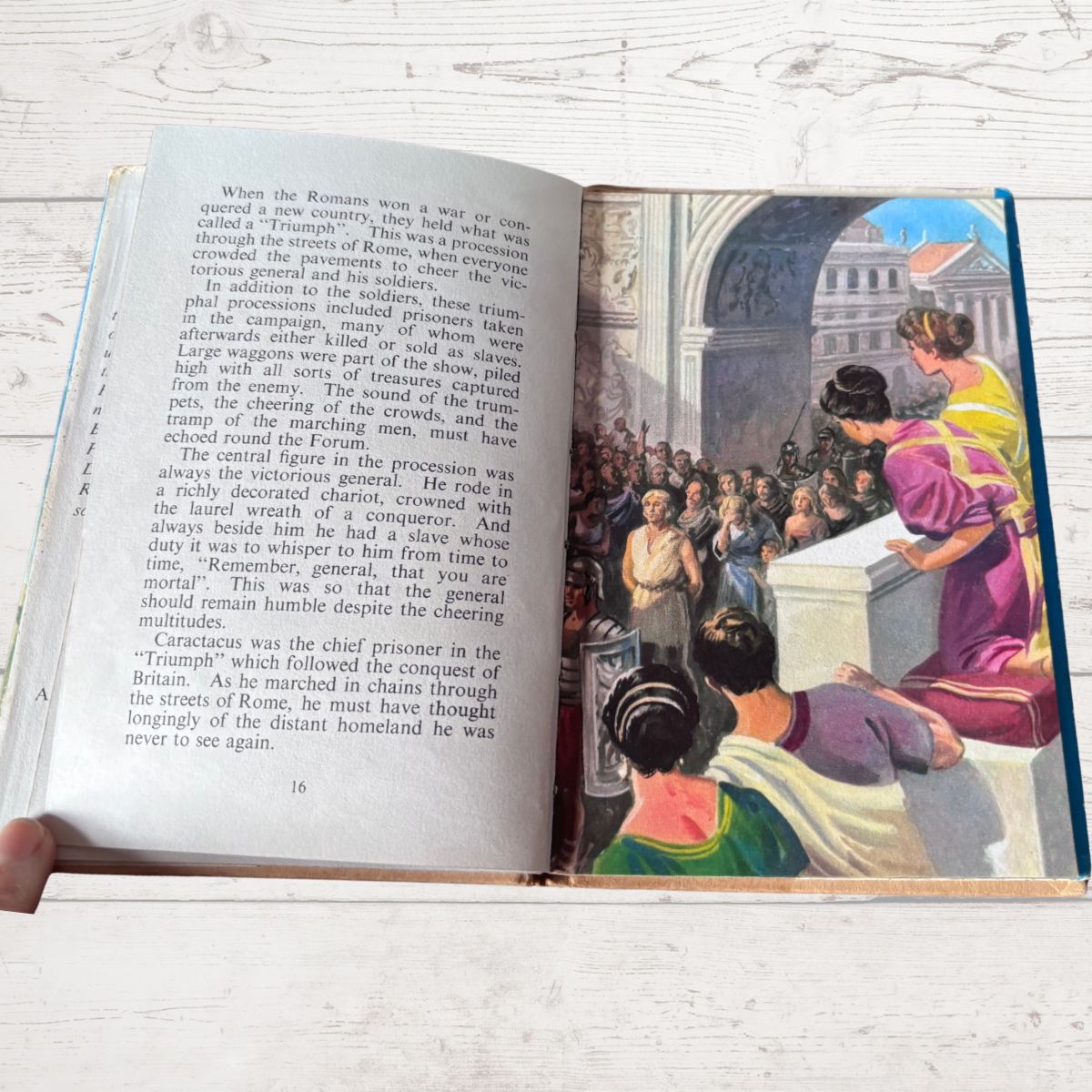 An open children's book lying on a light wood surface, showing printed text on the left page and a colorful illustrated scene on the right page with a woman, child, and horse against a building background.