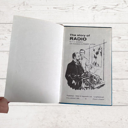 An open vintage book titled 'The story of RADIO' held open by a hand, showing a black-and-white illustration on the right page against a white wood-plank background.