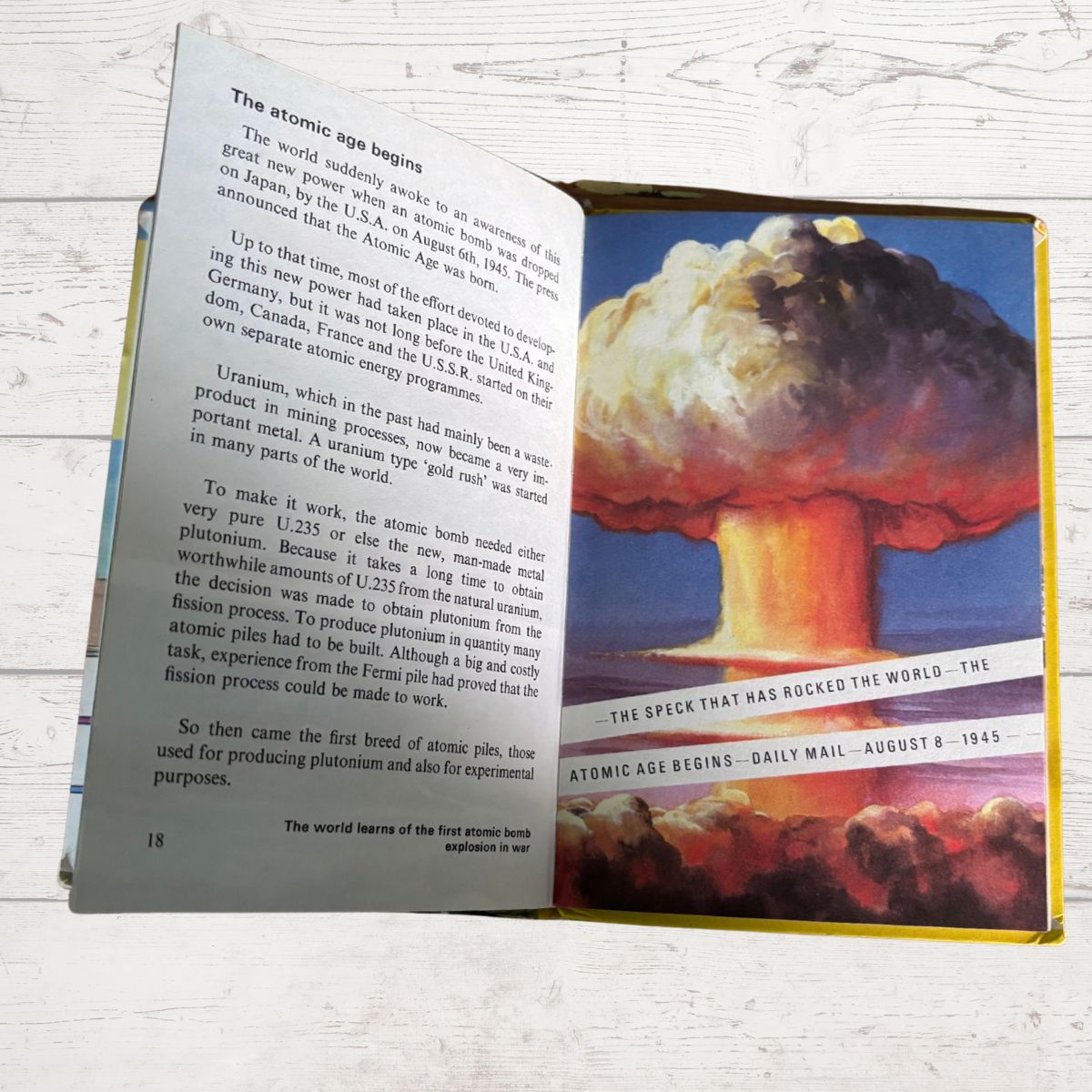 An open book showing printed pages about the atomic age and a large cover or spread with a dramatic red, orange, and yellow abstract design and the text 'The Spook That Was the World: The Atomic Age Begins - Daily Mail - August 1945.'.