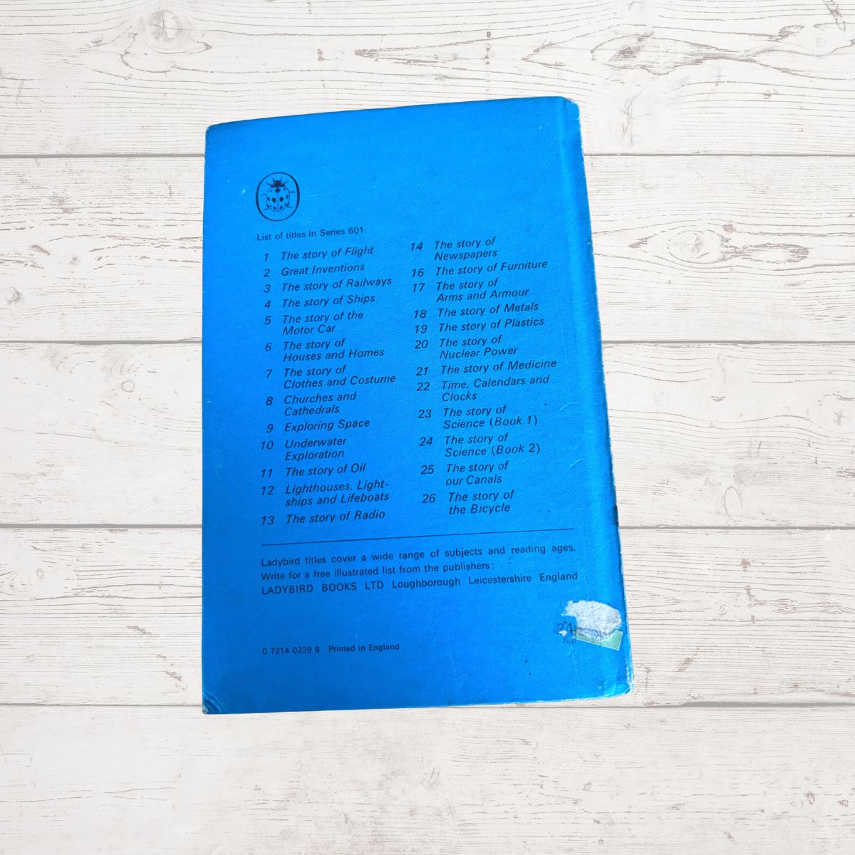 A blue hardcover book or booklet lying on a white wood surface, showing the back cover with printed tracklist-style text and a small circular logo near the top left.