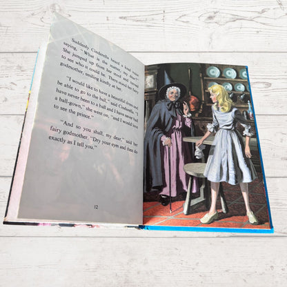 An open children's book lying on a white wood surface, showing printed text on one page and a colorful illustration on the other. The illustration depicts an elderly woman in a dark cloak and a young girl in a light blue dress in an indoor scene.