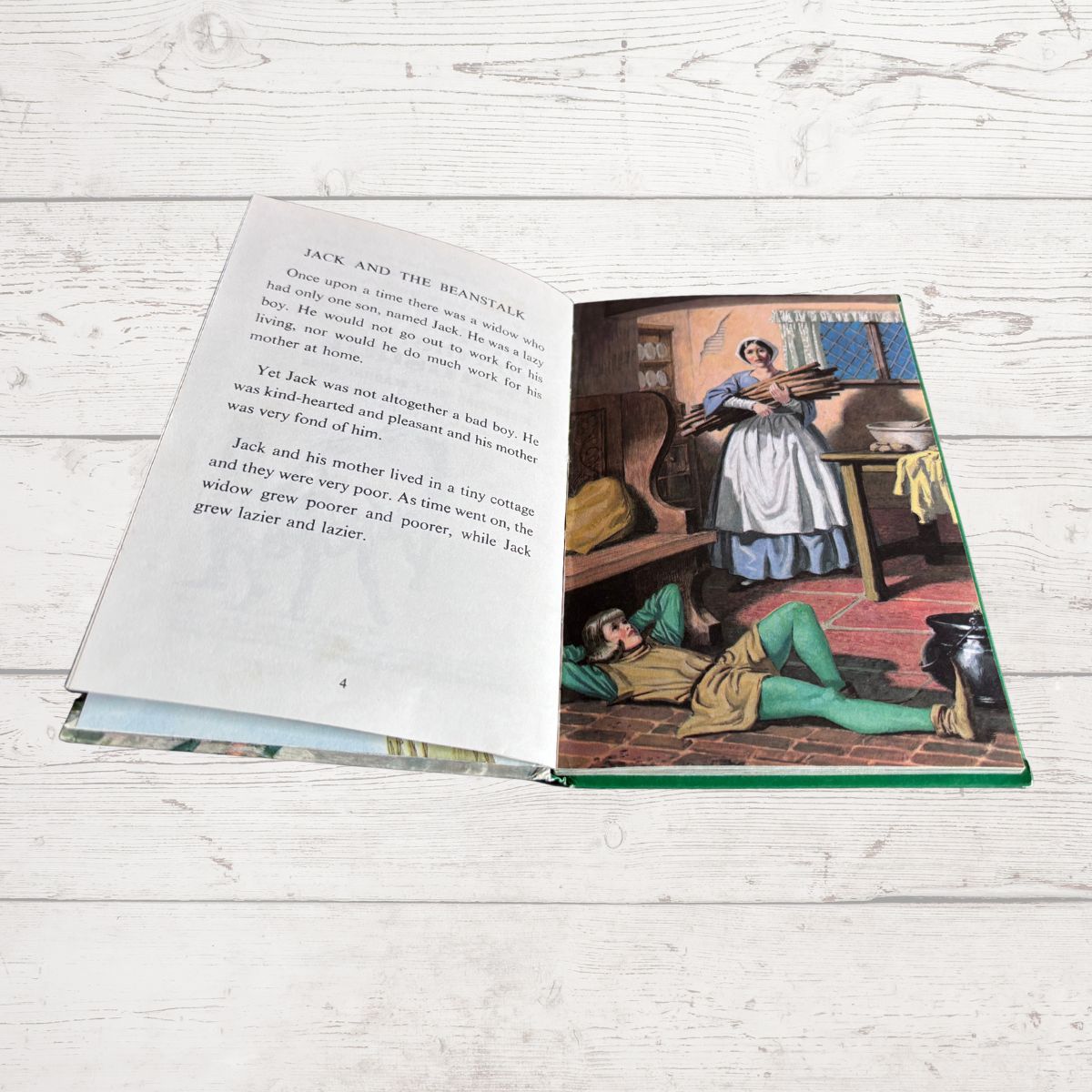 An open children's book lying on a white wood plank surface, showing a printed story page with text on the left and a colorful illustration on the right of a woman holding a basket while another person lies on the floor.