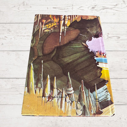 A vintage illustrated book featuring a surreal illustration of a cave with spikes and a large creature, displayed on a light gray wood plank background