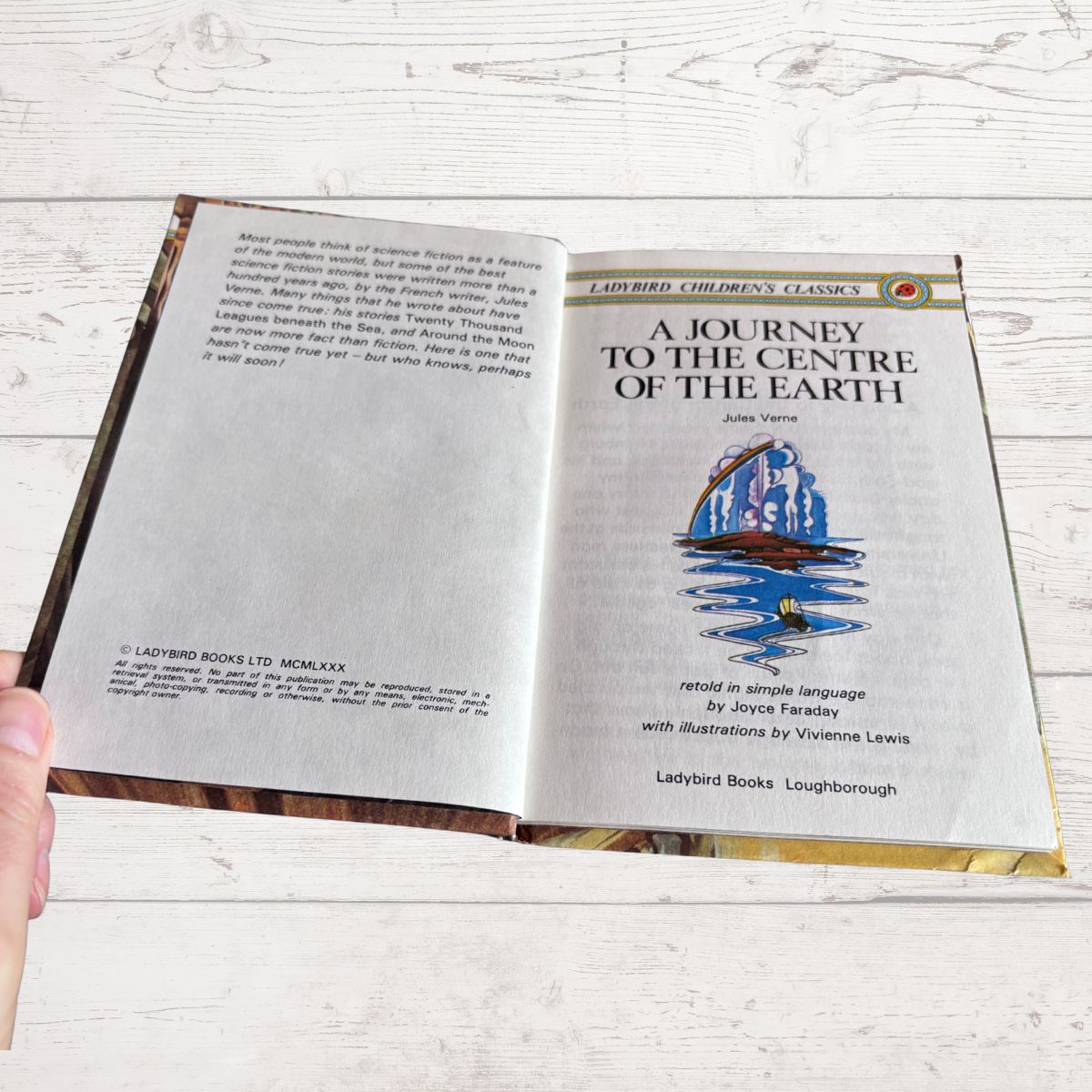 An open hardcover book titled 'A Journey to the Centre of the Earth' held open by a hand, showing the illustrated cover page on the right and the title page on the left, set against a light wood background.