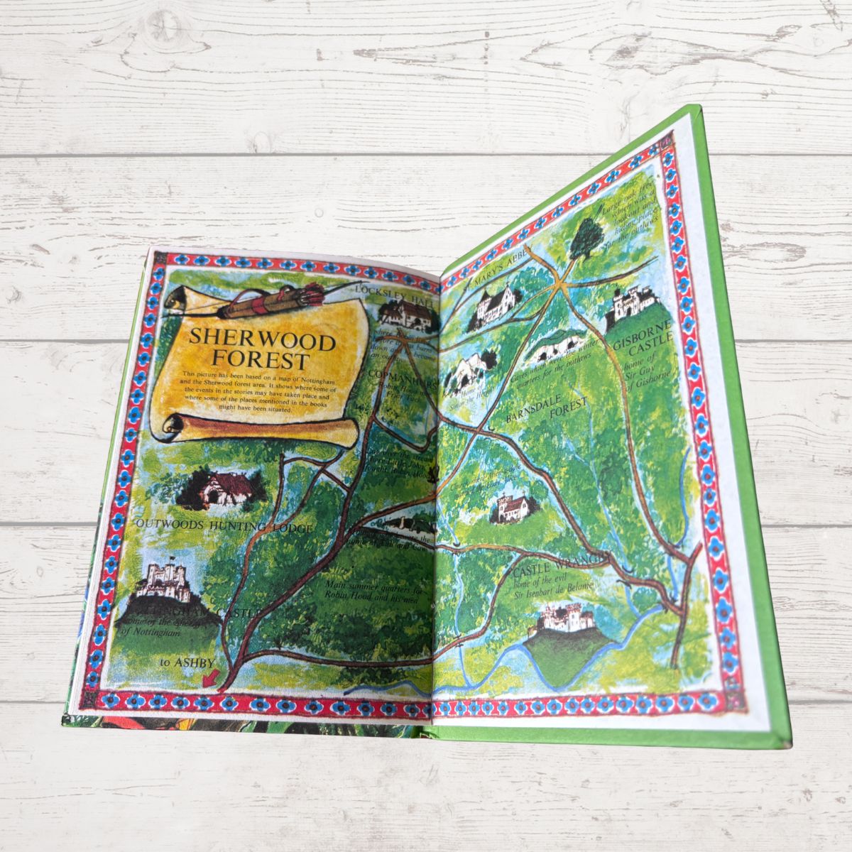 An open illustrated book or map titled 'Sherwood Forest' laid flat on a light whitewashed wood surface. The pages show a colorful fantasy-style forest map with green hills, trees, houses, and decorative borders.