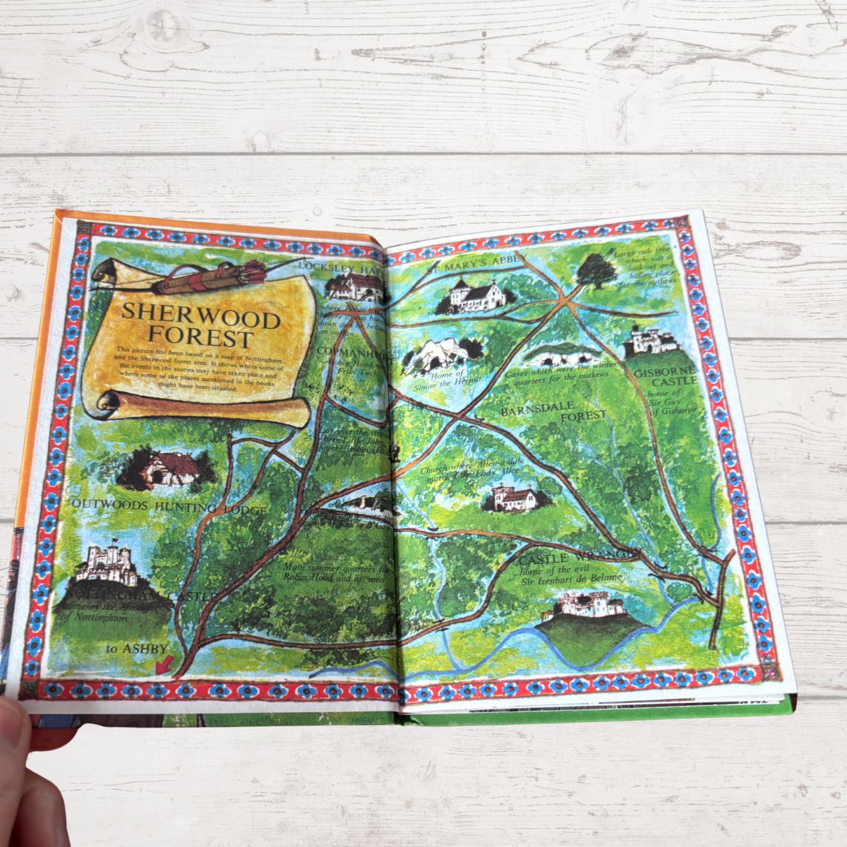 An open illustrated map book titled 'Sherwood Forest' laid on a white wood surface, with a hand holding one corner. The pages show a colorful fantasy-style map with green forest, winding brown paths, blue water, and small castle and house illustrations.