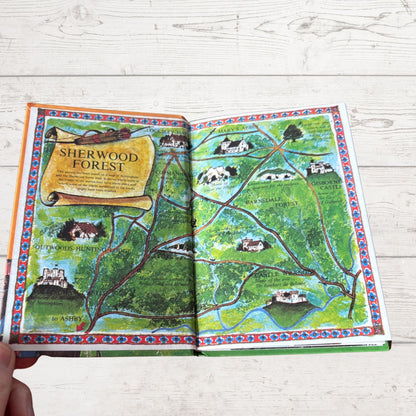 An open illustrated map book titled 'Sherwood Forest' laid on a white wood surface, with a hand holding one corner. The pages show a colorful fantasy-style map with green forest, winding brown paths, blue water, and small castle and house illustrations.