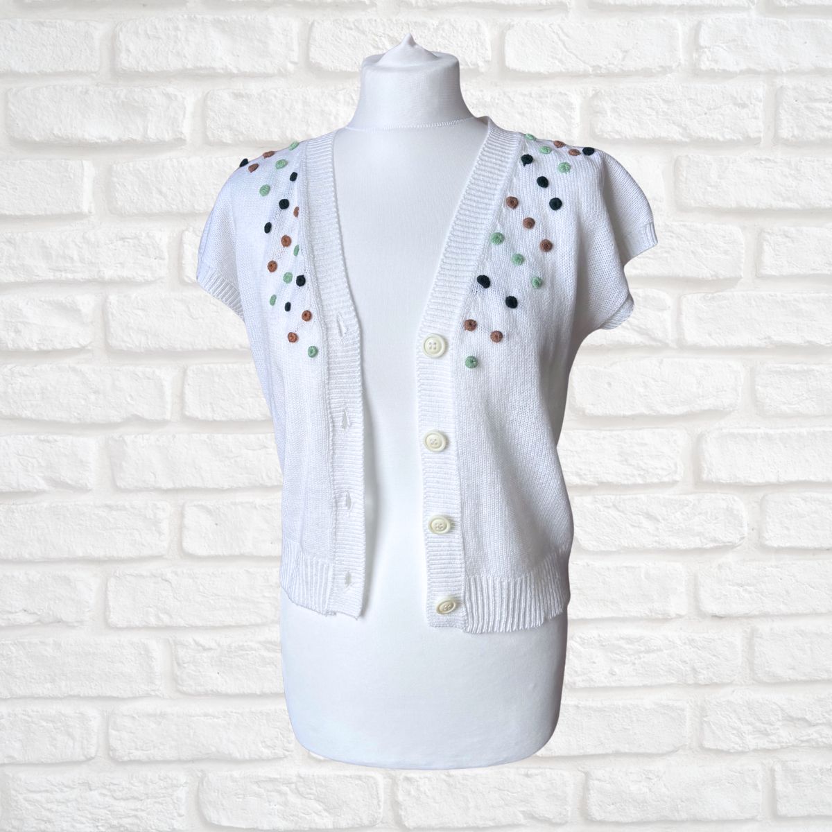 A white short-sleeve knit cardigan sweater displayed on a white mannequin torso, featuring decorative multicolored pom-poms along the shoulders and front buttons, set against a white brick wall background.