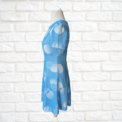 Blue dress with white geometric patterns on a mannequin against a white brick wall.
