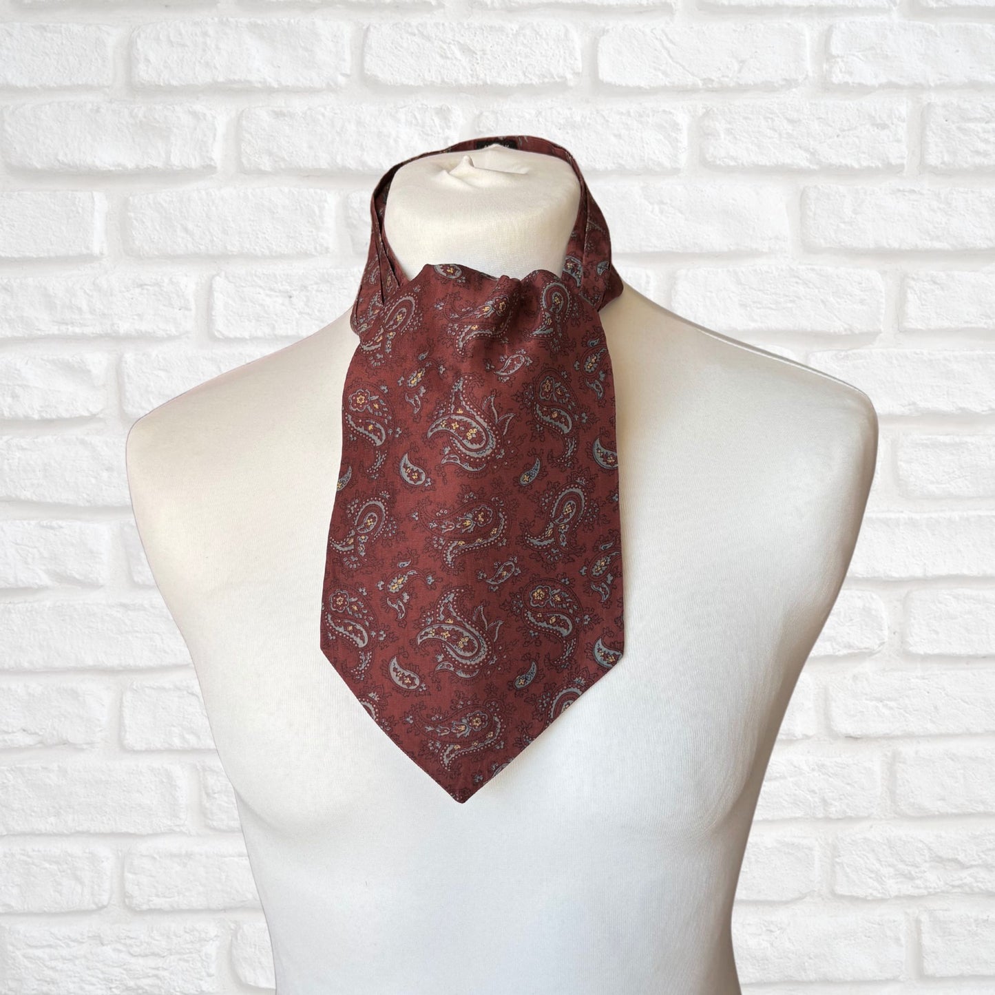 A vintage silk cravat featuring a paisley print in red, grey, yellow, and blue, displayed on a mannequin against a white brick wall background.