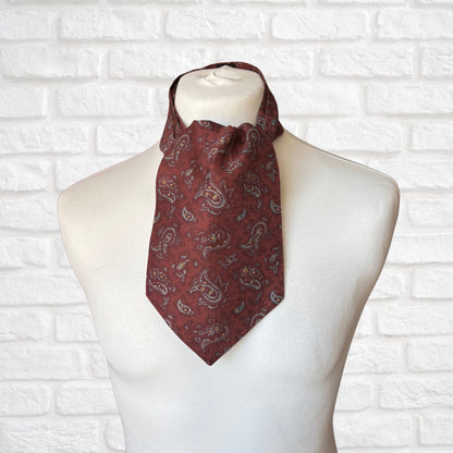 A vintage silk cravat featuring a paisley print in red, grey, yellow, and blue, displayed on a mannequin against a white brick wall background.