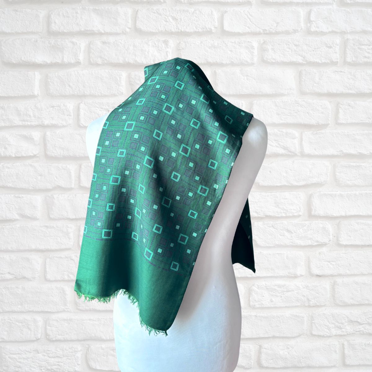 A green patterned scarf draped on a white mannequin torso, set against a white brick wall background.