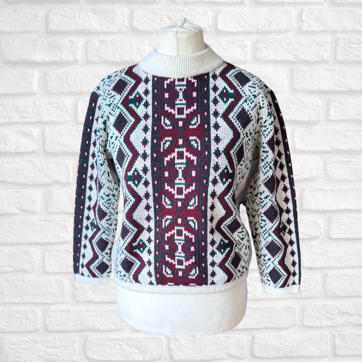 A patterned knit sweater displayed on a white mannequin torso against a white brick wall. The sweater features a white ribbed mock neck and cuffs, with a geometric Fair Isle style pattern in burgundy, navy blue, and white.