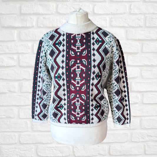 A patterned knit sweater displayed on a white mannequin torso against a white brick wall. The sweater features a white ribbed mock neck and cuffs, with a geometric Fair Isle style pattern in burgundy, navy blue, and white.