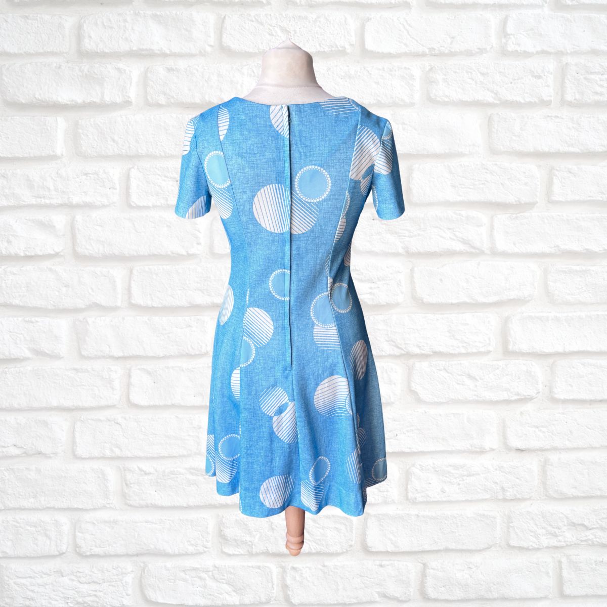 Blue dress with white circular pattern on a mannequin against a white brick wall.