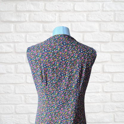 A sleeveless dress displayed on a light blue mannequin torso, featuring a dense multicolour floral print on a dark navy base, set against a white brick wall background.