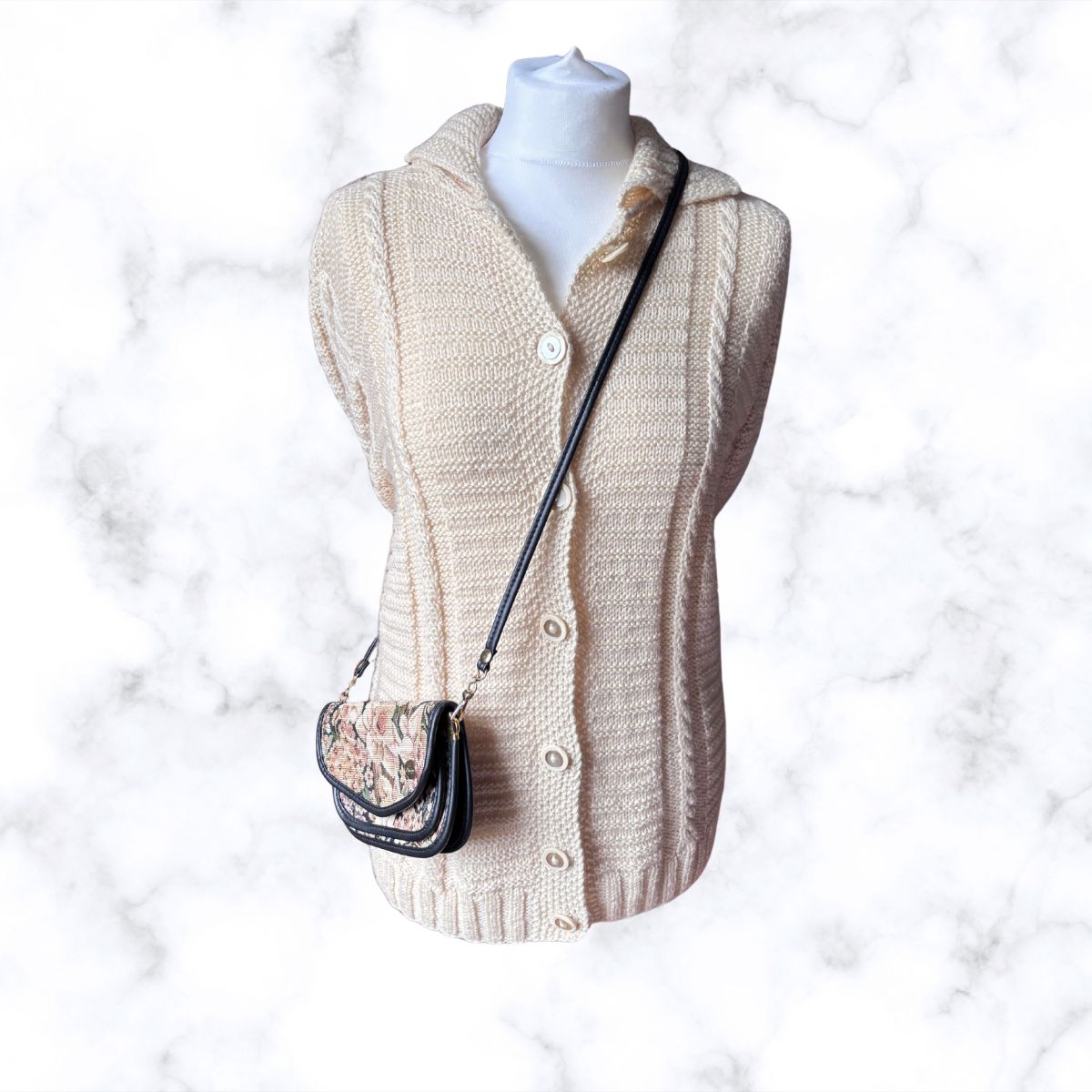 A cream cable-knit cardigan sweater displayed on a white mannequin torso, styled with a small black shoulder bag featuring a floral patterned front and black strap, set against a white marble background.