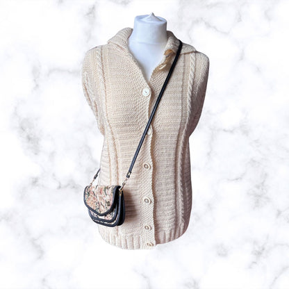 A cream cable-knit cardigan sweater displayed on a white mannequin torso, styled with a small black shoulder bag featuring a floral patterned front and black strap, set against a white marble background.