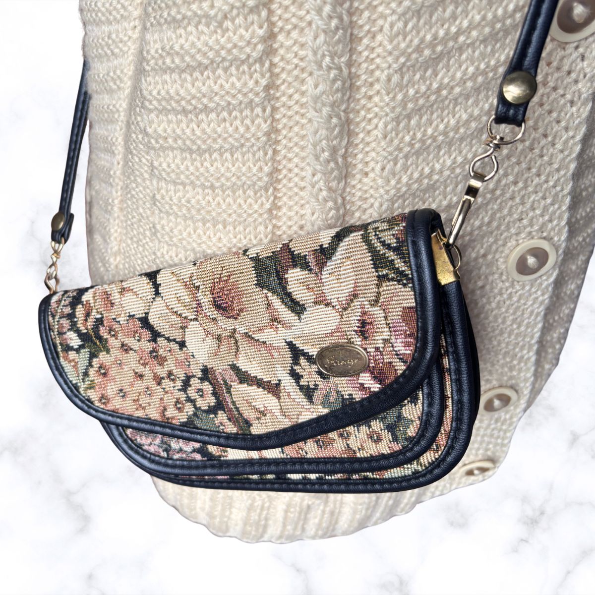 A close-up of a small handbag with a floral tapestry pattern and dark navy trim, shown resting against a cream knit sweater on a light marble surface.
