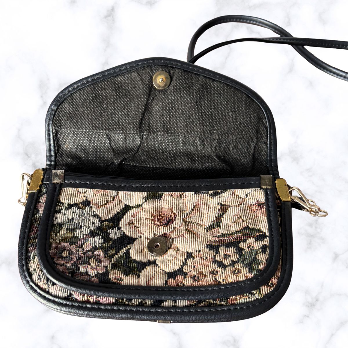 A vintage-style crossbody handbag with a black leather trim and strap, featuring a floral tapestry patterned front pocket and a black mesh interior, displayed open on a white marble background.