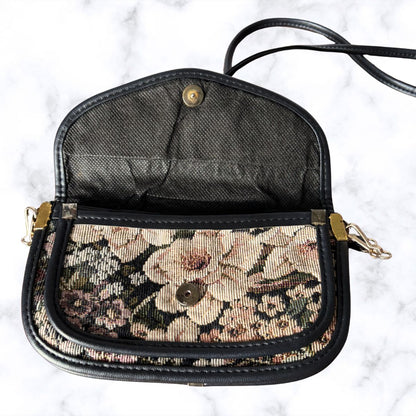 A vintage-style crossbody handbag with a black leather trim and strap, featuring a floral tapestry patterned front pocket and a black mesh interior, displayed open on a white marble background.