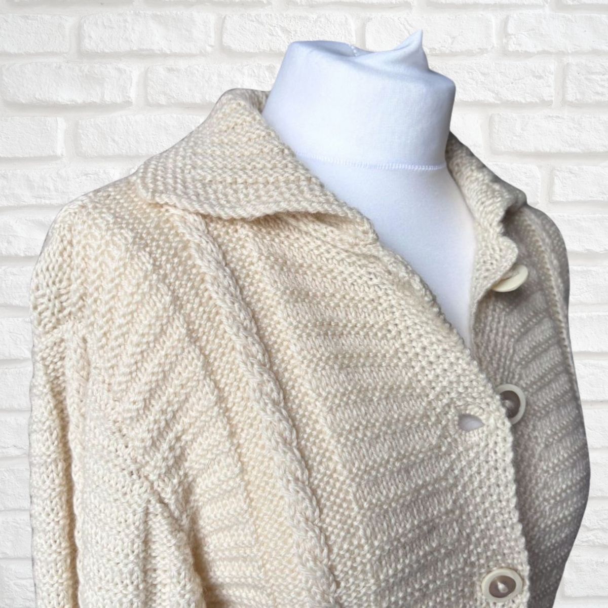 A cream-colored knit cardigan sweater with a textured herringbone pattern, displayed on a white mannequin torso against a white brick wall background.