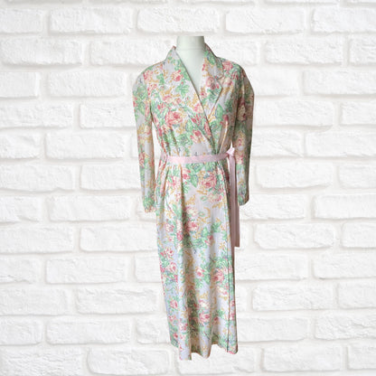 A vintage 80s full-length pink rose print dressing gown with a pink ribbon belt, displayed on a mannequin against a white brick wall.
