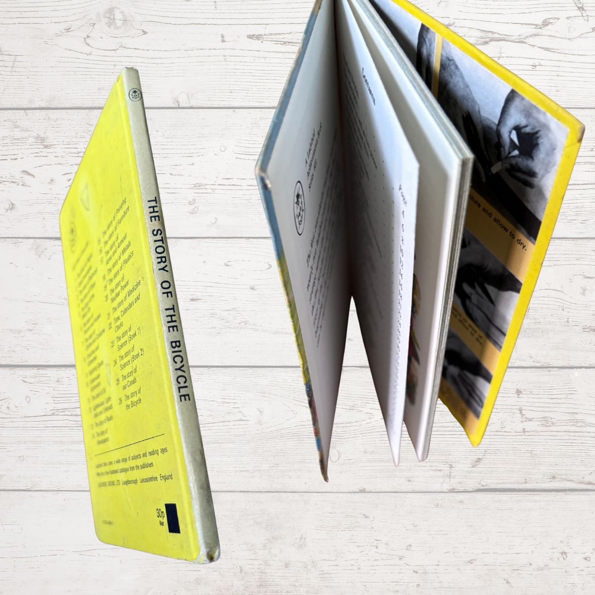 A book titled 'The Story of the Bicycle' shown standing upright with its back cover visible, alongside an open view showing multiple interior pages and a bright yellow page edge, set against a light wood plank background.